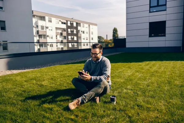 An urban businessman sits on a grass in front of the business center and hanging on social media.