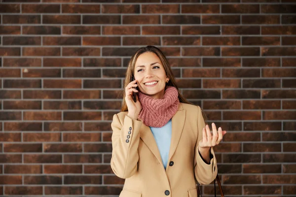 A woman standing on the street and talking on the phone. A happy fashionable young woman in warm clothes standing on the street in front of the brick wall and talking on the phone.
