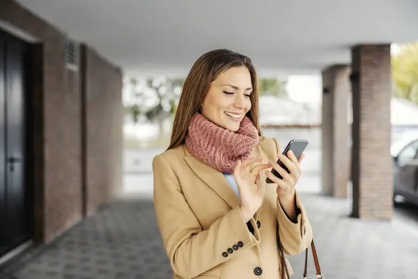 Telecommunications worldwide and technology. A happy woman in a coat is standing outdoors and typing a message on her smartphone she holding in her hands.