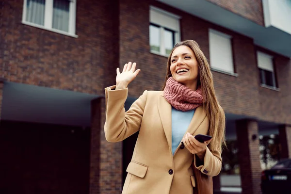 A woman wave on the street, greeting concept. A young happy stylish woman in a warm outfit standing on the street with a phone in her hands and greets the neighbors.