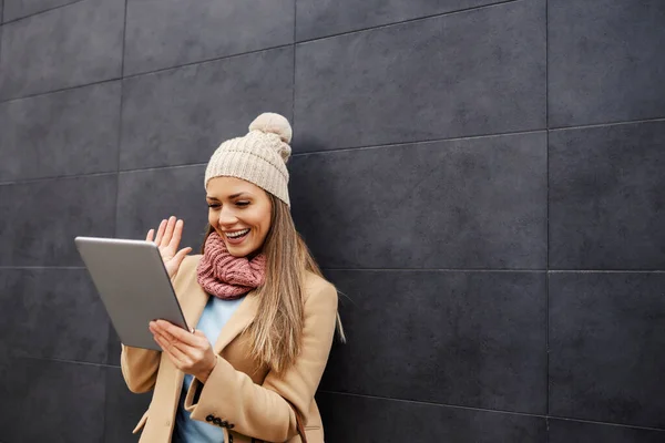社交媒体平台和网络通信 一个穿着保暖衣服的年轻而快乐的女人靠着墙壁 在平板电脑上播放视频 挥挥手 — 图库照片