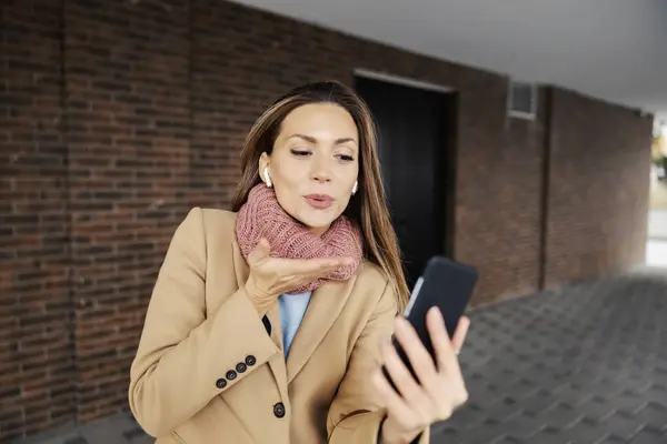 Telecommunications, internet, and social media apps. A young woman in a warm outfit standing outdoors with wireless headphones, having a video call, and sending a kiss.