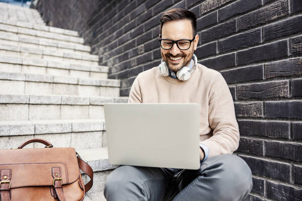 A smart casual man sits on the stairs outside and smiling at the laptop while typing on it. — Stock Image Smart Casual Man Sits Stairs Smiling Laptop While Typing Stock Image