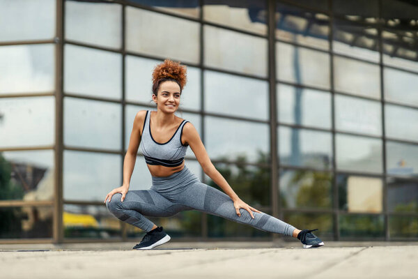 A happy flexible sportswoman stretching her legs and warming up before running in an urban district. An urban sportswoman warming up outdoors. — Stock Image Happy Flexible Sportswoman Stretching Her Legs Warming Running Urban District Stock Picture
