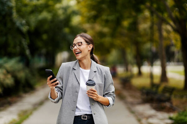 A happy young businesswoman is laughing and holding a cup of coffee while looking at the phone and standing in a park. A woman found something funny on her phone. A businesswoman in a park on a coffee break — Stock Image Happy Young Businesswoman Laughing Holding Cup Coffee While Looking Phone Stock Picture