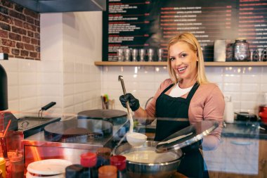 A pancake house worker is holding ladle with flapjacks mix and smiling at the camera. clipart