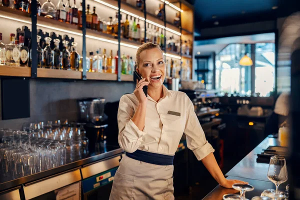 Portrait of a professional female bartender smiling while having a phone conversation on a break behind the counter in a bar.