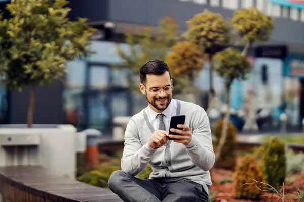 An elegant happy man is sitting on a bench in park and typing messages on the phone while smiling at it.