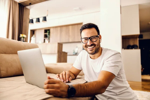A man sits at home and searching internet on laptop and smiles at the camera.