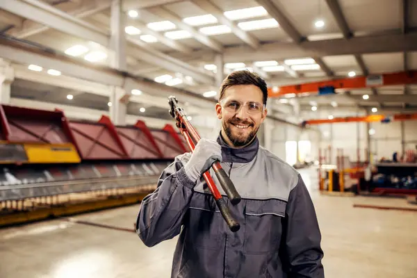 A metallurgy worker is holding industrial wire cutter on his shoulder and smiling at the camera on his way to workspace.