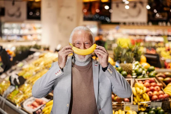 A goofy senior man is pretending banana is his mouth and standing at supermarket while looking at the camera.