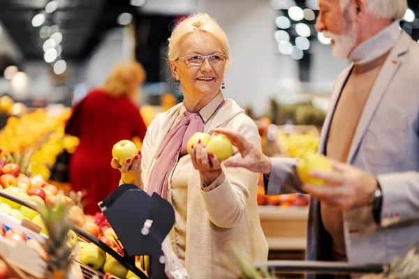 A cheerful pensioners are picking fresh apples at the supermarket and buying it.
