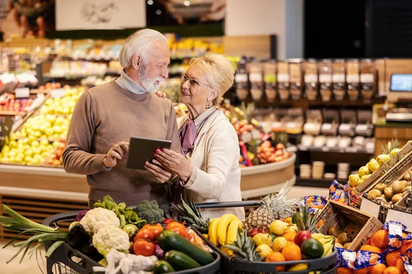 A happy old couple is buying groceries from list on tablet at the supermarket.