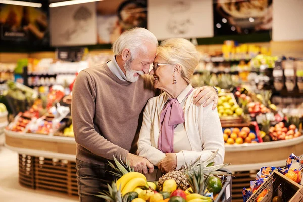 A cute old couple in love is embracing at the supermarket while purchasing groceries.