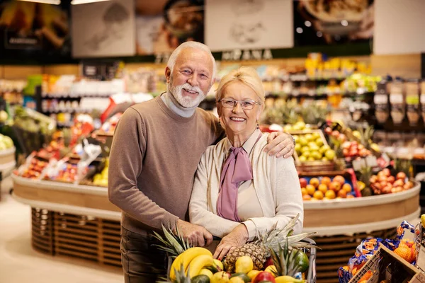 A cute old couple in love is hugging at the supermarket and purchasing while smiling at the camera.