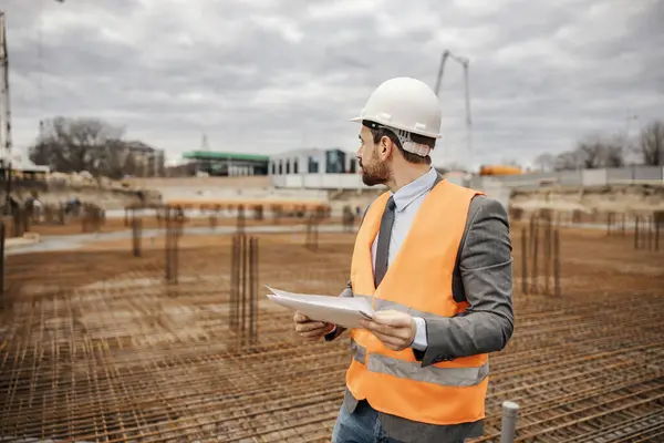 エンジニアが再建エリアに立って 彼の手にある計画と文書を持ち 建物の基礎に関する作品を見ています — ストック写真