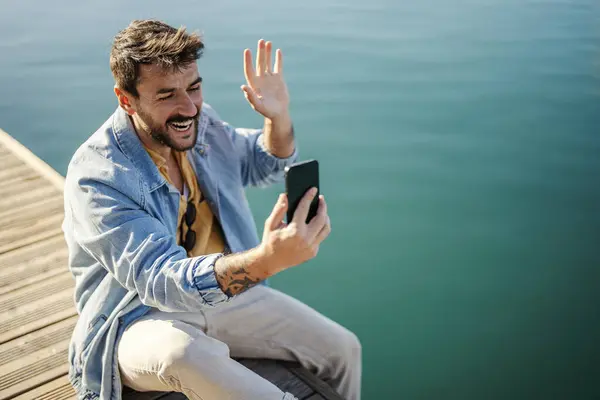 A happy man is greeting friends while sitting on the dock and having video call on the phone.