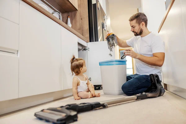 A father is emptying vacuuming cleaner into a garbage can while babysitting his baby girl.