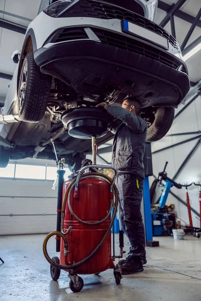 An auto-mechanic is draining oil from engine on a car while standing under the car at mechanic's shop.