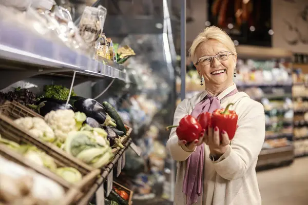 陽気なシニアの女性がスーパーマーケットに立ってカメラで微笑んでいる間に唐辛子を握っています — ストック写真