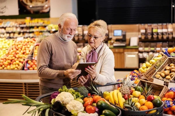A senior couple is looking and following groceries list on tablet at the supermarket.