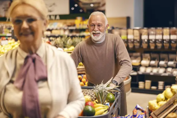 A happy senior man is pushing shopping cart full of groceries while purchasing in supermarket.