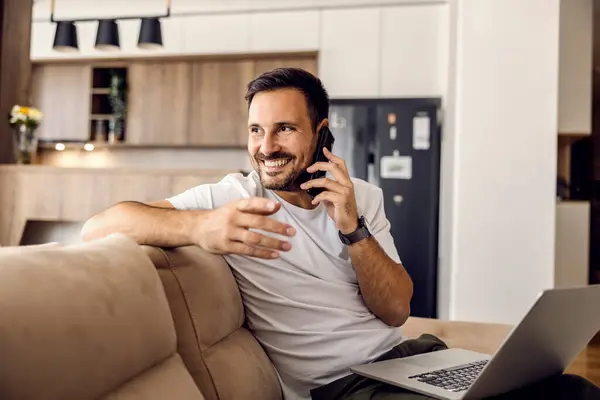 A happy man is having phone conversation while sitting at home on sofa with a laptop in his lap.