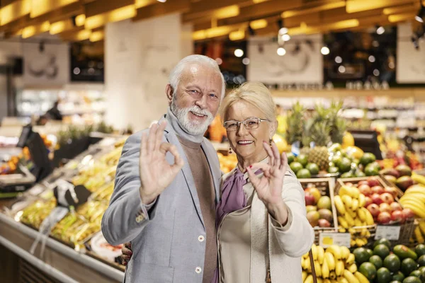 A cheerful old couple is gesturing ok sign while looking at the camera in supermarket.