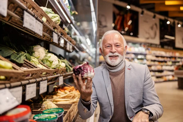 A happy old man is purchasing vegetables at the supermarket and smiling at the camera.