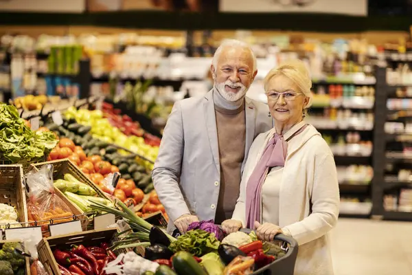 An old happy couple is pushing shopping cart full of vegetables while walking at the supermarket.