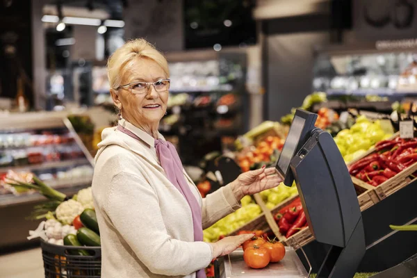 A happy old woman is measuring vegetables on scales at the supermarket while looking at the camera.