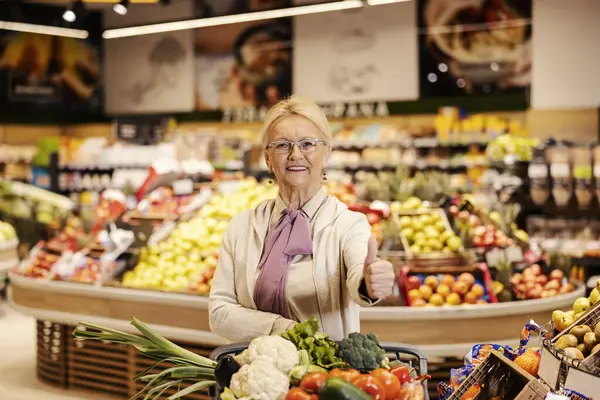 A happy old woman is pushing shopping cart at supermarket and giving thumbs up while smiling at the camera.