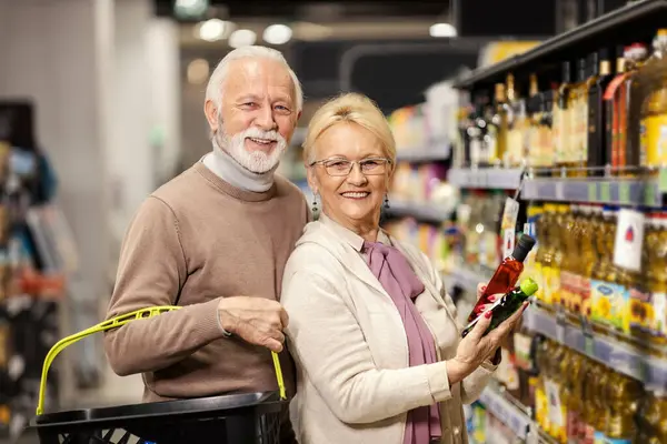 A cheerful senior couple is choosing oil at the supermarket while smiling at the camera.