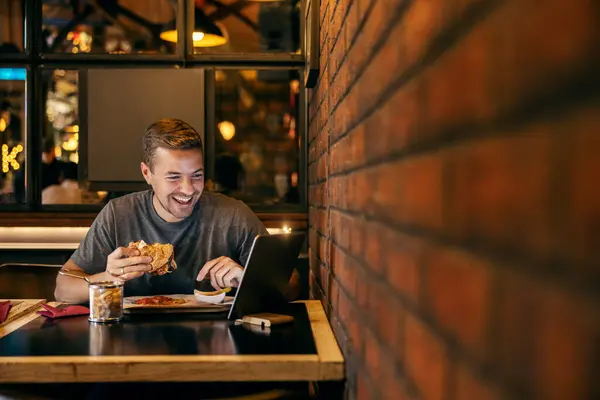 A happy man is eating hamburger at fast food restaurant and smiling at tablet.