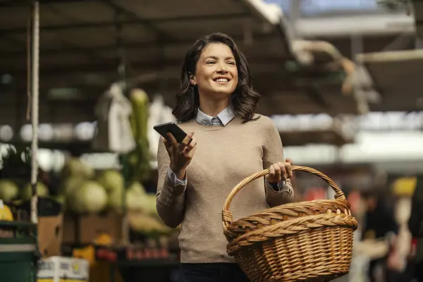 A happy woman with basket in hands is using a phone at marketplace while shopping.