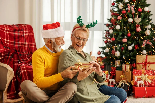 A surprised senior woman is receiving a christmas present from her husband on christmas and new year while celebrating it at home.