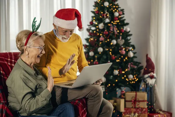 A happy grandparents sitting at home on christmas and new year and waving at laptop during video call.