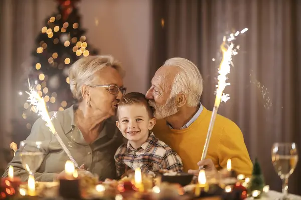 Affectionate grandparents celebrating Christmas and new year\'s eve at home with grandchild and kissing him while holding fireworks.