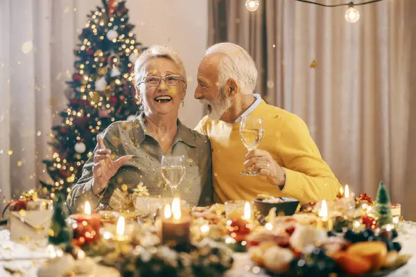 A festive excited grandparents celebrating christmas and new year\'s eve with confetti and hugging at home at dining table.