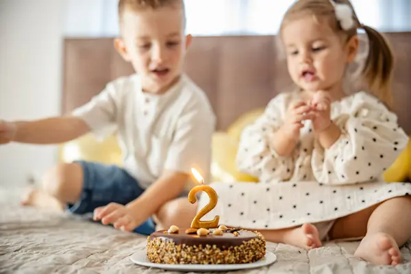 A brother and sister celebrating birthday while sitting with cake on a bed.