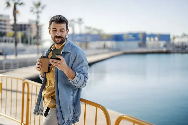 A happy retro man is standing at the dock with his phone and enjoying his coffee.