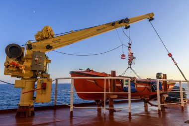 Turuncu kurtarma botu büyük kargo gemisi ve vinçte. Tatbikatta bir adam var. Cankurtaran botu eğitimi.