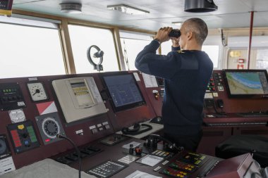 Seyir köprüsünde dürbünle nöbet tutan bir subay var. Mavi üniformalı beyaz adam kargo gemisinin köprüsünde..