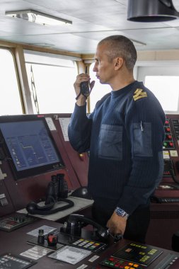 Seyir köprüsünde telsizle nöbet tutan bir subay var. Mavi üniformalı beyaz adam kargo gemisinin köprüsünde..