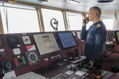 Seyir köprüsünde telsizle nöbet tutan bir subay var. Mavi üniformalı beyaz adam kargo gemisinin köprüsünde..