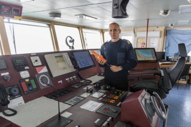 SART ile birlikte seyir köprüsünde nöbet tutan bir subay var. Mavi üniformalı beyaz adam kargo gemisi köprüsünde arama kurtarma radar vericisi kullanıyor..