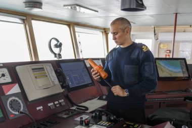 SART ile birlikte seyir köprüsünde nöbet tutan bir subay var. Mavi üniformalı beyaz adam kargo gemisi köprüsünde arama kurtarma radar vericisi kullanıyor..