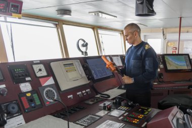 SART ile birlikte seyir köprüsünde nöbet tutan bir subay var. Mavi üniformalı beyaz adam kargo gemisi köprüsünde arama kurtarma radar vericisi kullanıyor..