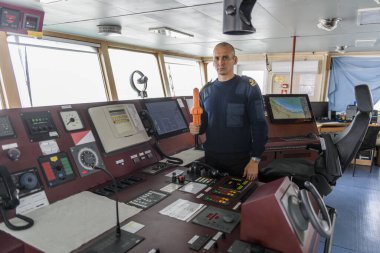 SART ile birlikte seyir köprüsünde nöbet tutan bir subay var. Mavi üniformalı beyaz adam kargo gemisi köprüsünde arama kurtarma radar vericisi kullanıyor..