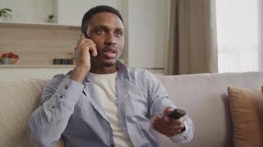 TV izlerken telefonla konuşan genç 20 'li yaşlarda Afro-Amerikan bir erkek uzaktan kumandasını kullanırken kablo ağıyla sohbet ediyor.